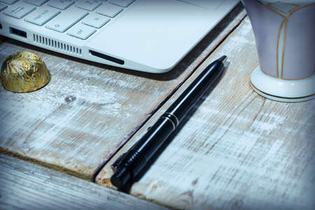 Laptop, pen, coffee cup and candy in a golden wrapper on a wooden table with traces of white paintの写真素材