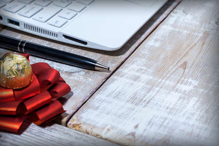 Laptop, pen, candy in a gold wrapper and a red bow on a wooden table with traces of white paintの写真素材