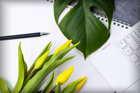 A large green leaf, a bunch of yellow tulips, a white notebook, pen and spiral notebook on a gray...