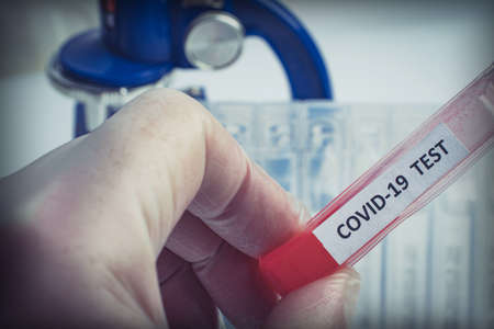 A hand in a protective glove holds a test tube labeled COVID-19 with red liquid against the background of a microscope and a number of clean medical tubes.の写真素材