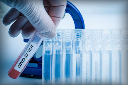 A hand in a protective glove holds a test tube labeled COVID-19 with a blood test against a...
