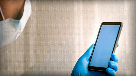 A man in a medical mask holds a smartphone with a white blank screen in his hand in a blue medical glove.の写真素材