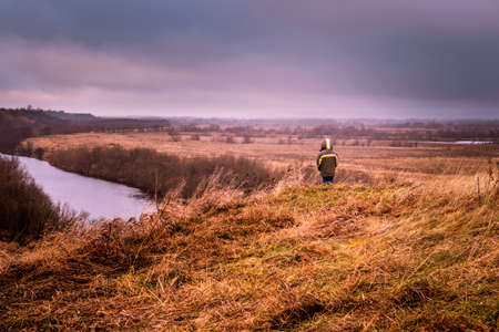 A young man stands on the top of a hill overlooking the river valley against the backdrop of a...