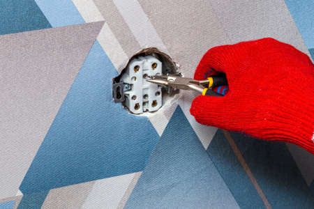 An electrician wearing red protective gloves uses a screwdriver to install a socket into the wall of the living room. Close-up of hands. Home master concept.の写真素材