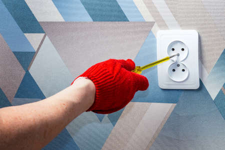 An electrician wearing red protective gloves uses a screwdriver to install a socket into the wall of...