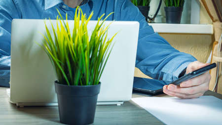A young man with a phone in his hand works at a white laptop at home at a table near the window. Home office, copy spaceの写真素材