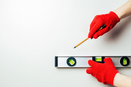 The hands of a worker in red protective gloves hold a hammer and a nail against a white wall. Home master conceptの写真素材