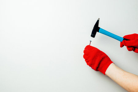 The hands of a worker in red protective gloves hold a hammer and a nail against a white wall. Home master conceptの写真素材