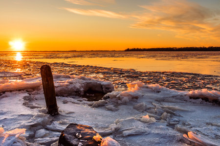 A view of a river covered in ice illuminated by the orange light of the sun at sunset. Winter landscapeの写真素材