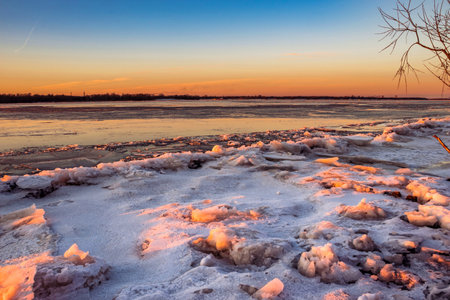 A view of a river covered in ice illuminated by the orange light of the sun at sunset. Winter landscapeの写真素材