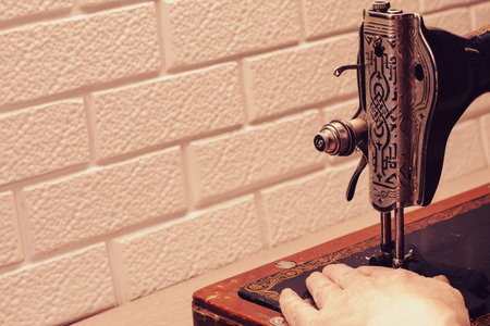 A hand advances the fabric in an old sewing machine with a wooden table against a white brick wall....