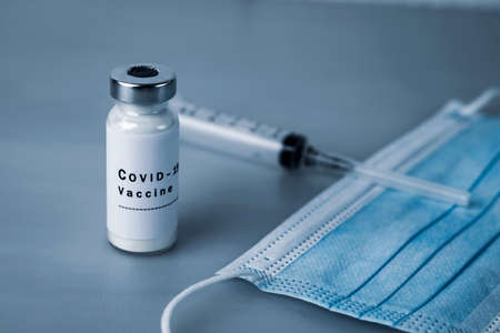 Vaccine ampoule with COVID-19 inscription, injection syringe and medical protective mask on a gray background. Coronavirus vaccination conceptの写真素材