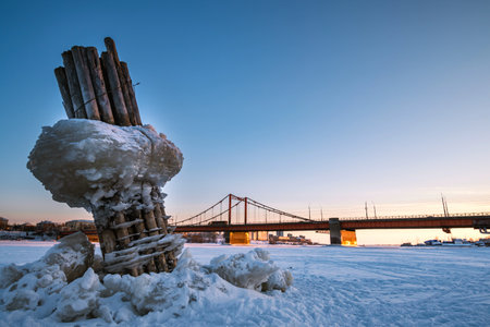 Wooden pillars with blocks of ice stick out from under the ice against the background of the bridge over the river.の写真素材
