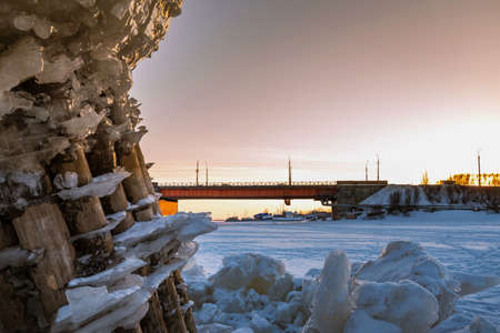 Wooden pillars with blocks of ice stick out from under the ice against the background of the bridge over the river.の写真素材