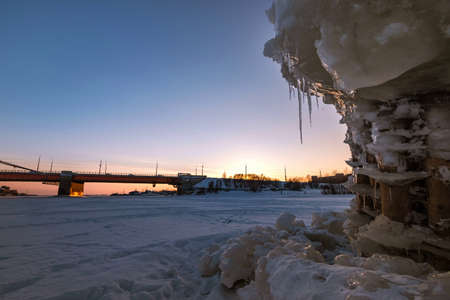 Wooden pillars with blocks of ice stick out from under the ice against the background of the bridge over the river.の写真素材