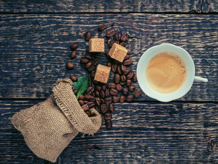 A cup of frothy coffee, a bag of spilled coffee beans and cane sugar lumps on an old wooden surface....