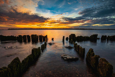 Beautiful sunset in the sky with clouds over the river. Lines of old wooden supports from the pier in the water in the foreground. Landscape at sunsetの写真素材