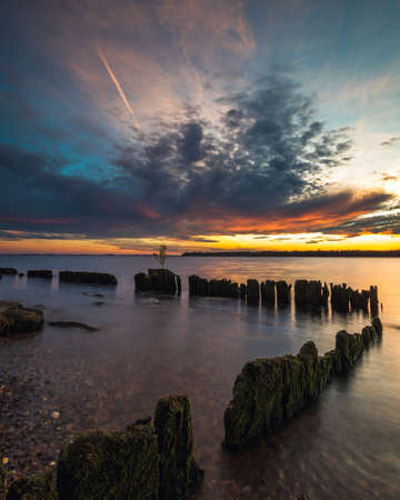 Beautiful sunset in the sky with clouds over the river. Lines of old wooden supports from the pier in the water in the foreground. Landscape at sunsetの写真素材