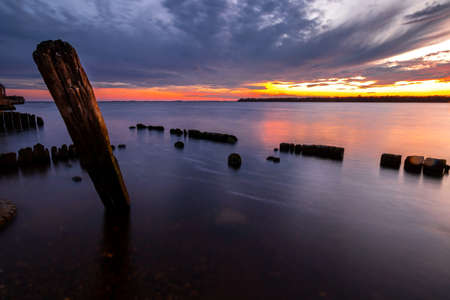 Beautiful sunset in the sky with clouds over the river. Lines of old wooden supports from the pier in the water in the foreground. Landscape at sunsetの写真素材