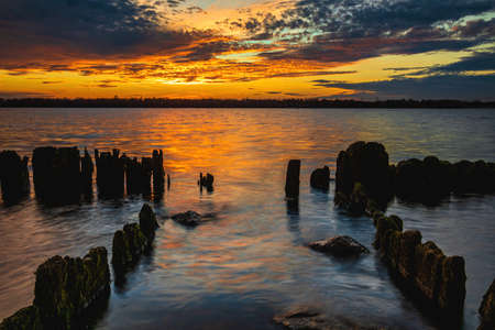 Beautiful sunset in the sky with clouds over the river. Lines of old wooden supports from the pier in the water in the foreground. Landscape at sunsetの写真素材