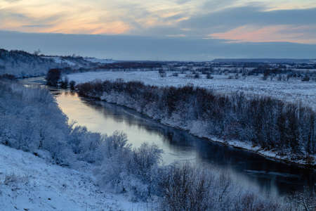 A beautiful winter view from the hill to the river with frosty trees growing along the banks. Winter landscape at sunsetの写真素材