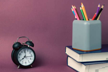 Black alarm clock, a stack of books and a gray glass of office supplies with multicolored pencils on a purple background. Back to school conceptの写真素材