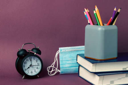 Black alarm clock, a stack of books, multicolored pencils in a glass and medical masks on a purple background. Coronavirus restrictionsの写真素材