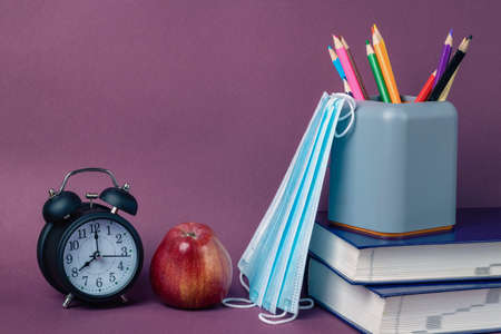 Black alarm clock, red apple, medical masks and school supplies on a purple background. Back to school, coronavirus restrictionsの写真素材