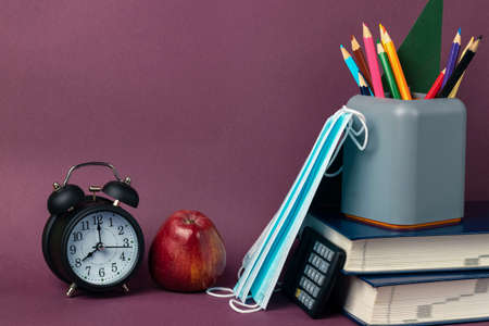Black alarm clock, red apple, medical masks and school supplies on a purple background. Back to school, coronavirus restrictionsの写真素材