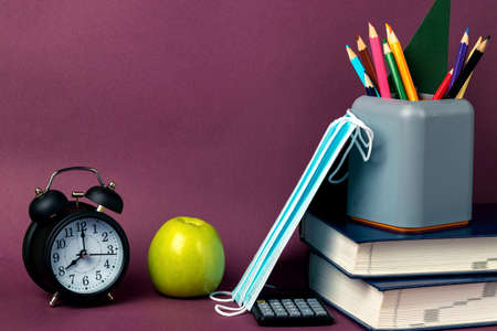 Black alarm clock, green apple, medical masks and school supplies on a purple background. Back to school, coronavirus restrictionsの写真素材