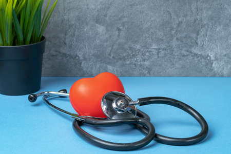 Stethoscope and rubber red heart on a blue table with a plant against a gray marble wall.の写真素材