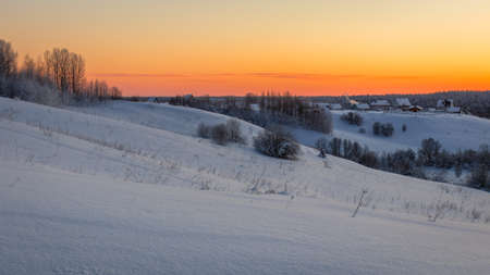 View of hills covered in snow with sparse trees and a small village against an orange sky.の写真素材