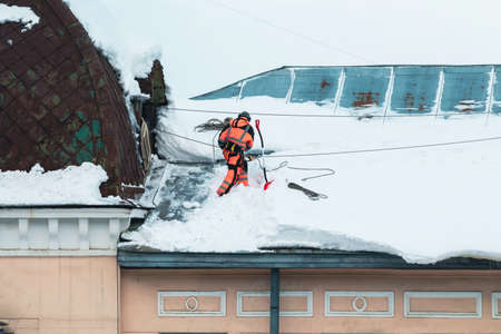 A working man in bright overalls with a safety belt with a shovel clears snow from the roof of an old building. Prevention of snow falling from the roof, industrial mountaineeringの写真素材