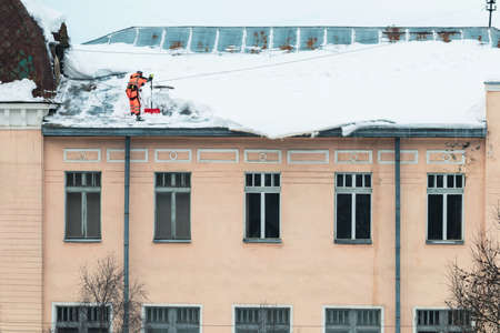 A working man in bright overalls with a safety belt with a shovel clears snow from the roof of an old building. Prevention of snow falling from the roof, industrial mountaineeringの写真素材
