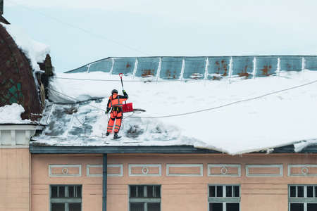 A working man in bright overalls with a safety belt with a shovel clears snow from the roof of an old building. Prevention of snow falling from the roof, industrial mountaineeringの写真素材
