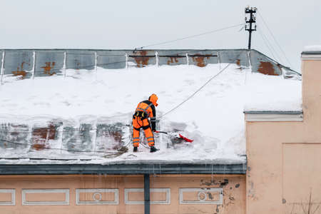 A working man in bright overalls with a safety belt with a shovel clears snow from the roof of an old building. Prevention of snow falling from the roof, industrial mountaineeringの写真素材