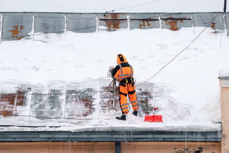 A working man in bright overalls with a safety belt with a shovel clears snow from the roof of an old building. Prevention of snow falling from the roof, industrial mountaineeringの写真素材