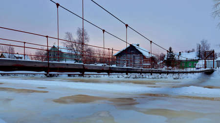 Suspension pedestrian bridge over a frozen river with wooden houses on the shore. rural landscapeの写真素材
