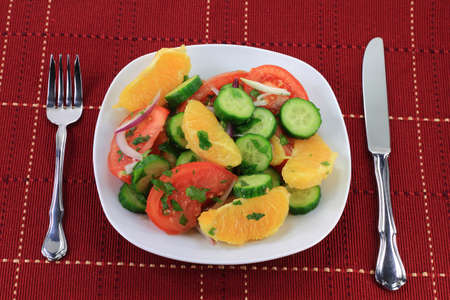 Salad  Vegetables - cucumbers, tomatoes, Fruit,orange, Seasoning - red onion, cilantro, Dressing  lime juice, olive oil in white porcelain dish and silverware of fork, knife over red serviette  の写真素材