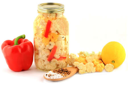 Florets Orange and White Cauliflower for pickling with lemon, pieces sliced Red Pepper in a glass jar, Pickling Salt and Seasoning in wooden cooking spoon, over white background  の写真素材