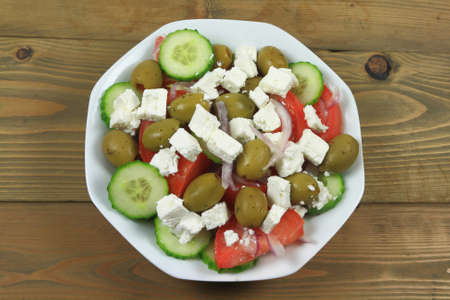 Greek salad from fresh organic vegetables: chopped red onion, tomatoes cut on wedges, sliced seedless cucumber; and stuffed green colossal olives, cubes goat Feta Cheese in white dish on wooden tableの写真素材