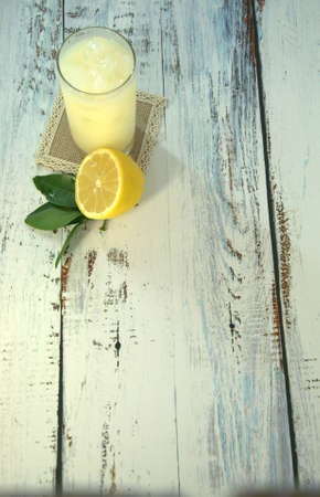A glass cup with a refreshing juice and ice, on a textile stand and cut half of a lemon with leaves lies on a white wooden table. Close-up.の写真素材
