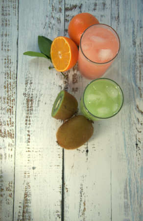 Two glass glasses with a refreshing juice and ice, on a textile stand, whole and sliced half of an orange with leaves and fruit, lies on a white wooden table. Close-up.の写真素材