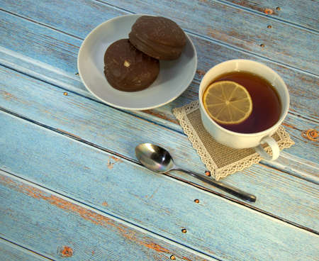 A cup of tea with lemon, a spoon and two chocolate cakes on a plate lie on a wooden table. Close-up.の写真素材