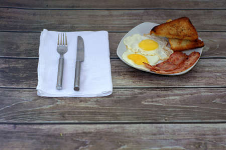Ceramic plate with fried bacon and eggs with croutons, lie on the table at the napkin with a fork and knife.の写真素材