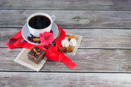 A cup of black coffee on a saucer decorated with spices, flowers and a scarlet ribbon, next to a napkin with a gift box. Close-up.の写真素材