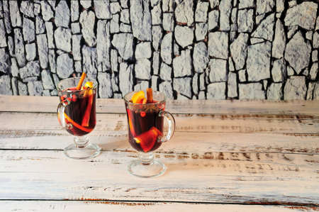 Two glasses of hot wine with slices of fruit and spices, stand on a wooden table against a stone wall. Close-up.の写真素材