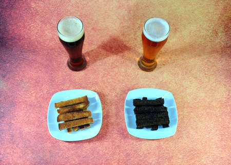 Two tall glasses with light and dark beer and two white ceramic plates with wheat and rye croutons, on a brown abstract background. Close-up.の写真素材