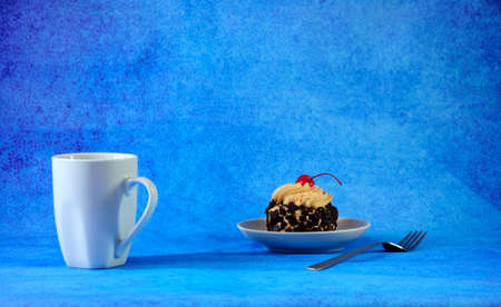A biscuit cake with chocolate chips and cherries on a cream hat on a white ceramic plate and a cup of black tea on a blue background. Close-up.の写真素材