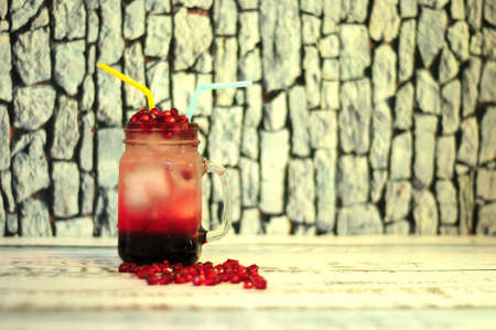 A mug of pomegranate juice with fruit grains and two straws stands on a wooden table by a stone...